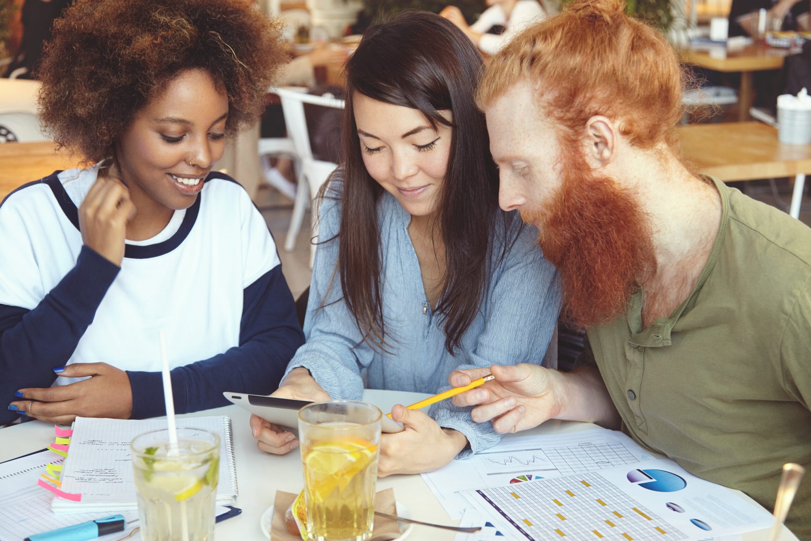 Multiethnic team of young ambitious entrepreneurs working together on start up project at cafeteria at table with papers of diagrams. Redhead bearded man pointing at touch pad screen using pencil