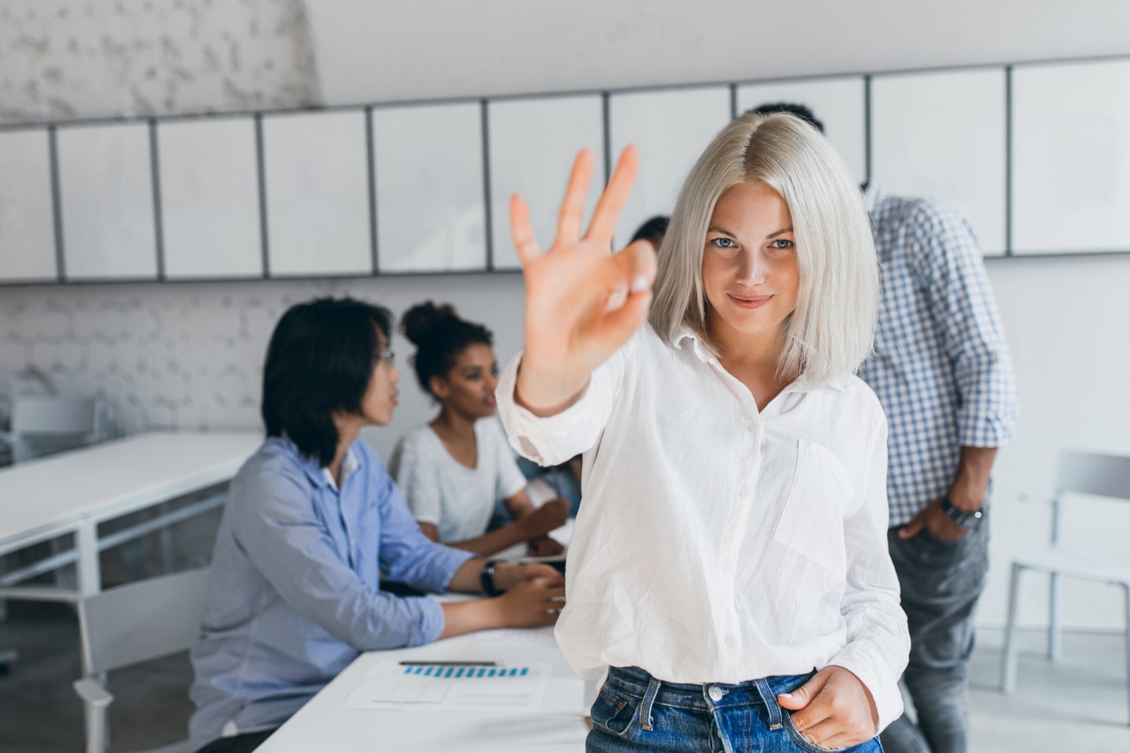 Slim girl with blonde short hair showing okay sign after succesful negotiation. Portrait of asian office worker posing with african colleague during conference with fair-haired woman on foreground.