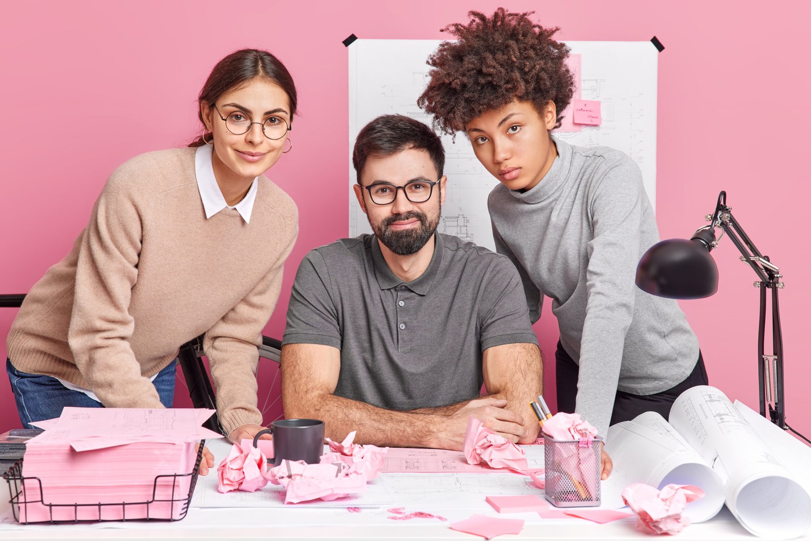 Three young motivated people discuss plan of cooperation on common architectural project poses in modern coworking office collaborate for making research isolated over pink background. Teamwork
