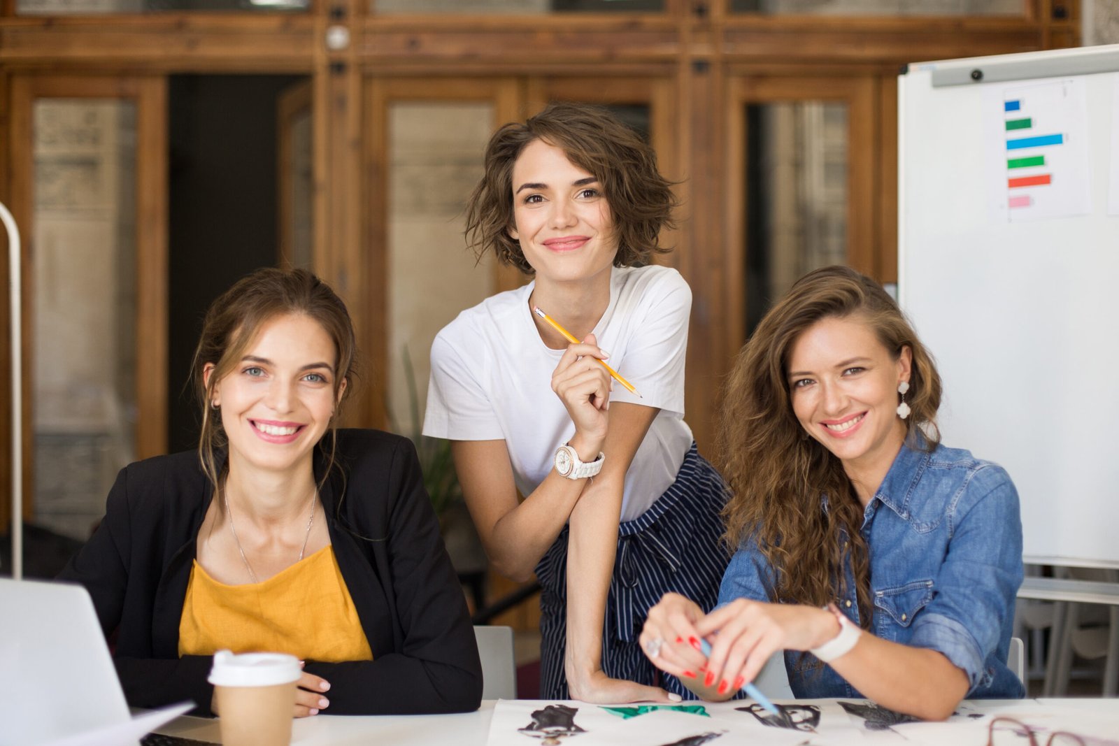 Young cheerful women happily looking in camera spending time at work. Pretty girls with laptop joyfully working with sketches together in modern cozy office